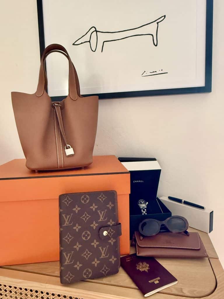 Luxury travel essentials for the Carry-On-Edit chapter: Hermès Picotin bag, Chanel J-12 watch, Céline sunglasses, Montblanc pen, Louis Vuitton agenda and passport.