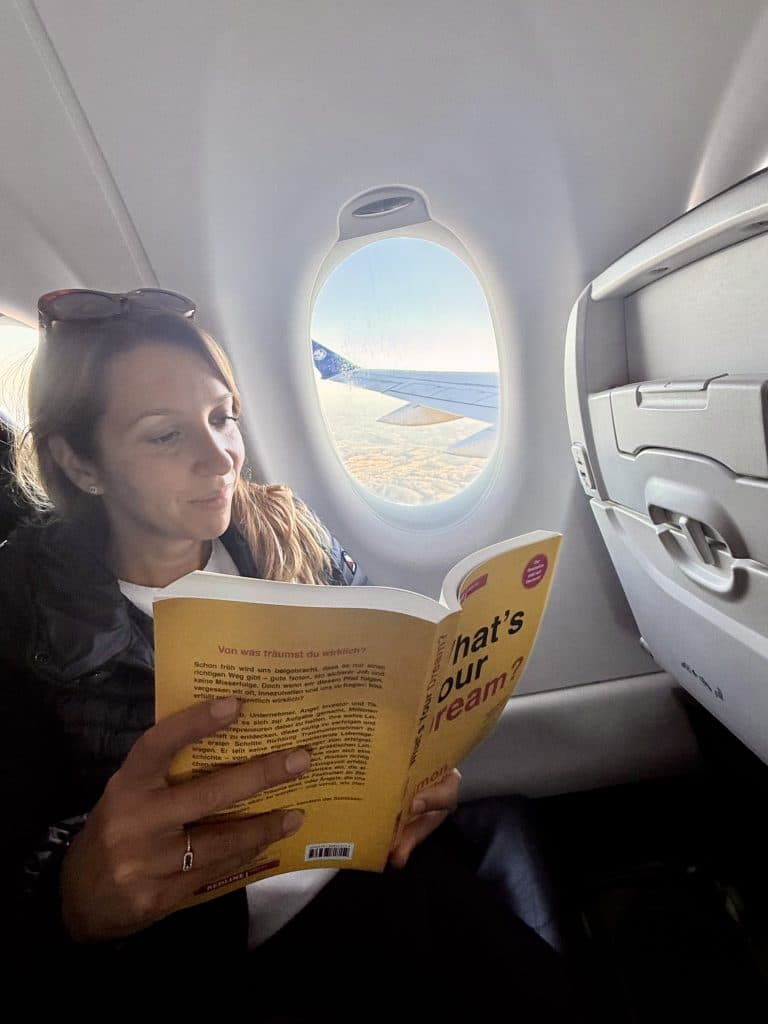 Woman reading the mindset book What's Your Dream on a plane, with a window view of the wing, symbolising luxury travel, lifestyle inspiration and reinvention at 40.